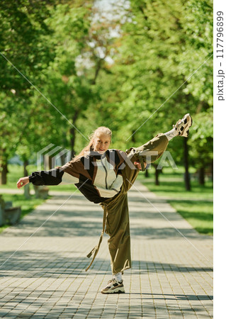 Person executing high kick balancing on one leg in serene urban park with green trees in background on sunny day wearing casual attire Person executing high kick balancing on one leg in serene urban park with green trees in background on sunny day wearing casual attire 117796899