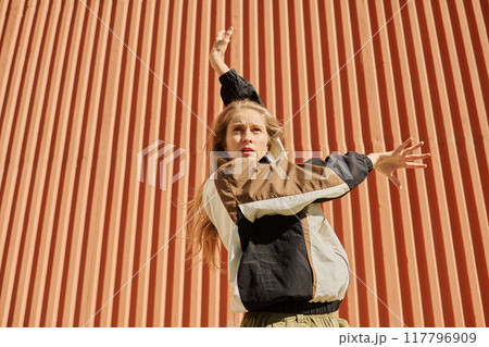 Blonde woman with long hair raising arm while posing against vertical striped wall wearing casual outfit and expressing artistic flair through dance movement Blonde woman with long hair raising arm while posing against vertical striped wall wearing casual outfit and expressing artistic flair through dance movement 117796909