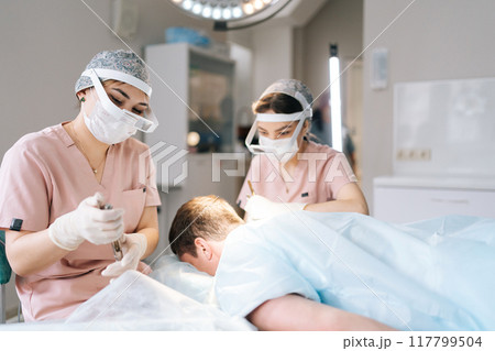 Two female trichologist performing local needle-free anesthesia to back of head using pistol in recipient area prior to hair transplant surgery in clinic. Concept of baldness treatment Two female trichologist performing local needle-free anesthesia to back of head using pistol in recipient area prior to hair transplant surgery in clinic. Concept of baldness treatment 117799504