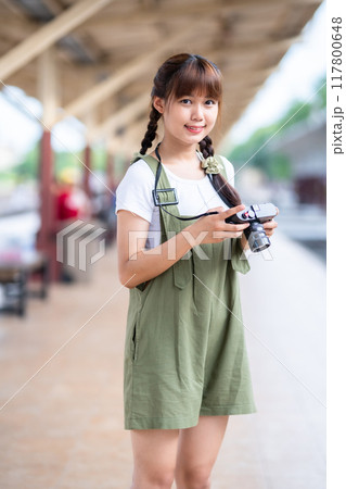 Portrait Young asian woman smiling tourist Traveler girl walking and with a holding the camera waits train travel journey is taken in railway platform Thailand, summer relax vacation Concept. 117800648