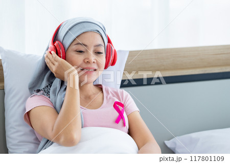 Happy a asian women disease mammary cancer patient with pink ribbon wearing headscarf in headphones is listening to music After treatment to chemotherapy sit on bed In the bedroom at the house 117801109