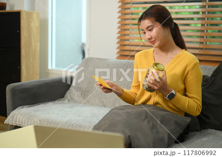 Happy young woman holding glass jar filled with coins and bank passbook sitting on sofa. Saving money concept 117806992