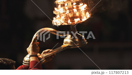 Varanasi, India. Man makes movements with a candle holder with lit candles on the Ganga Maha Aarti ceremony begins. Brahman priest wearing red and yellow dress doing traditional ganga aarti at ghat Varanasi, India. Man makes movements with a candle holder with lit candles on the Ganga Maha Aarti ceremony begins. Brahman priest wearing red and yellow dress doing traditional ganga aarti at ghat 117807213