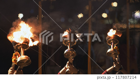 Varanasi, India. Man makes movements with a candle holder with lit candles on the Ganga Maha Aarti ceremony begins. Brahman priest wearing red and yellow dress doing traditional ganga aarti at ghat 117807214