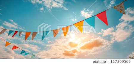 Colorful Festive Bunting Flags Against a Bright Blue Sky with Clouds and Sunlight 117809563