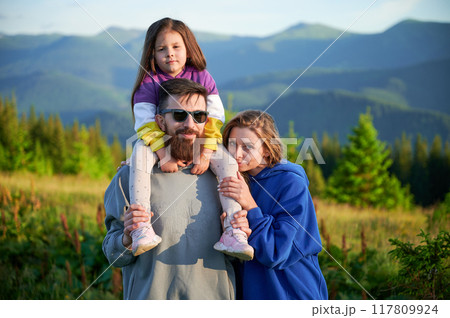 Family stands together in scenic meadow with backdrop of rolling mountains. Father carries his daughter on shoulders, both smiling and enjoying view, while mother stands beside them. 117809924