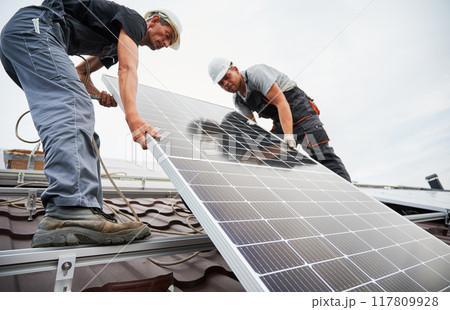Men installers lifting up photovoltaic solar moduls on roof of house. Electricians in helmets installing solar panel system outdoors. Concept of alternative and renewable energy. Men installers lifting up photovoltaic solar moduls on roof of house. Electricians in helmets installing solar panel system outdoors. Concept of alternative and renewable energy. 117809928
