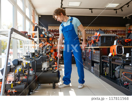 Young workman in overalls choosing pneumatic compressor at hardware store 117811568