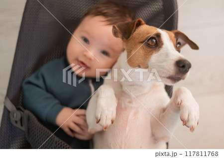 A dog and a cute three-month-old boy dressed in a blue overalls are sitting together in a baby lounger. A dog and a cute three-month-old boy dressed in a blue overalls are sitting together in a baby lounger. 117811768