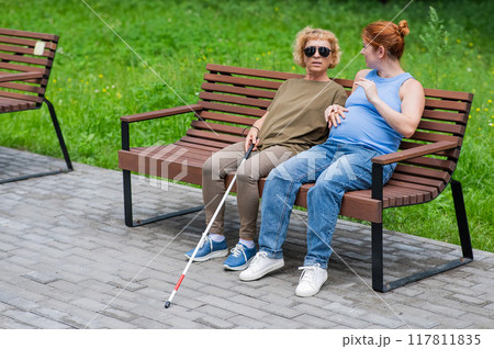 An elderly blind woman and her pregnant daughter are sitting on a bench in the park. An elderly blind woman and her pregnant daughter are sitting on a bench in the park. 117811835