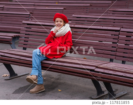 A pensive Caucasian girl in a red coat and beret sits on a bench.  117811855