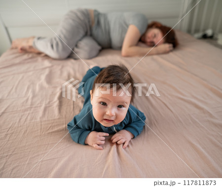 A three-month-old boy lies on his stomach on the bed and his mother sleeps behind him. Postpartum depression. A three-month-old boy lies on his stomach on the bed and his mother sleeps behind him. Postpartum depression. 117811873