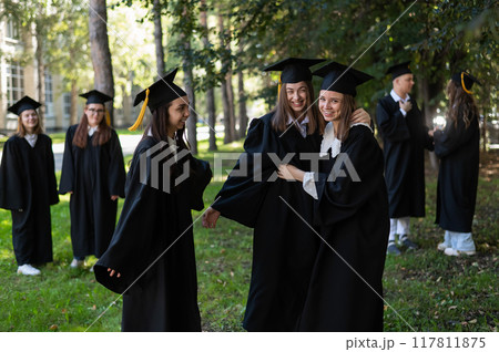 A group of graduates in robes congratulate each other on their graduation outdoors. A group of graduates in robes congratulate each other on their graduation outdoors. 117811875