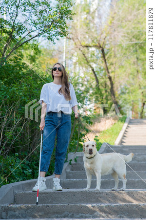 Young blind woman walking down the stairs in the park with a tactile cane and guide dog. Young blind woman walking down the stairs in the park with a tactile cane and guide dog. 117811879