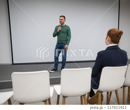 A red-haired Caucasian businesswoman sits in the front row of an empty conference room. Bearded man giving a lecture.  117811921