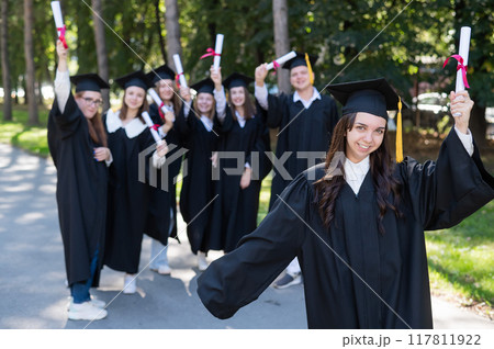 Group of happy students in graduation gowns outdoors. A young girl with a diploma in her hands in the foreground. 117811922