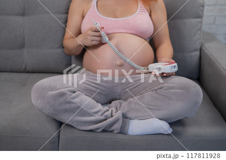 A pregnant woman uses a home doppler while sitting on the couch. Close-up of the abdomen. A pregnant woman uses a home doppler while sitting on the couch. Close-up of the abdomen. 117811928