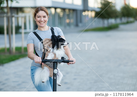 A woman rides an electric scooter with a dog in a backpack. Pappilion Spaniel Continental in a sling. 117811964