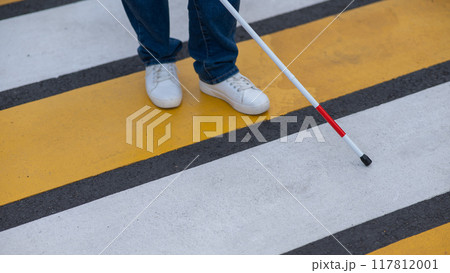 Close-up of the legs of a blind woman crossing the road at a crosswalk with a cane.  117812001