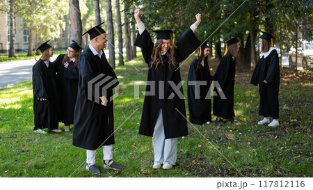 A group of graduates in robes congratulate each other on their graduation outdoors. 117812116