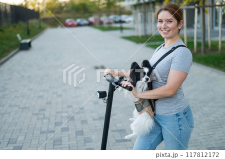 A woman rides an electric scooter with a dog in a backpack. Pappilion Spaniel Continental in a sling. 117812172