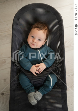 Cute newborn baby dressed in blue overalls sitting in a baby lounger. Vertical photo.  117812177