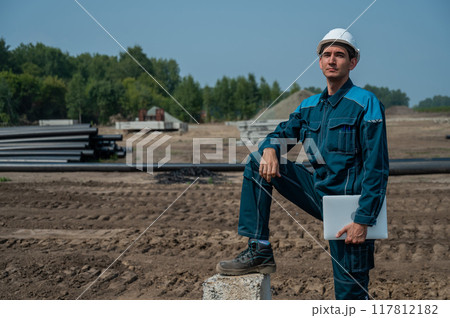 Caucasian male builder in hardhat holding laptop at construction site.  117812182