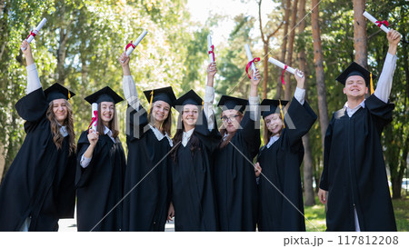 Row of young students in graduation gowns outdoors showing off their diplomas.  117812208