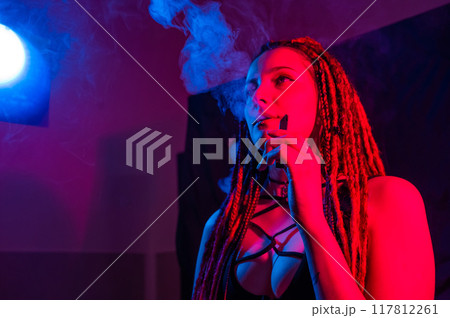 Caucasian girl with dreadlocks smokes a vape in red blue light. Caucasian girl with dreadlocks smokes a vape in red blue light. 117812261