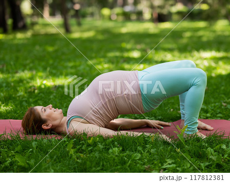 Pregnant caucasian woman doing yoga in the park. Prenatal shoulder bridge exercise.  117812381
