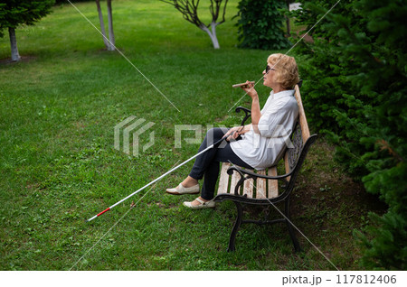 Elderly blind woman talking on a smartphone while sitting on a bench in the park. 117812406