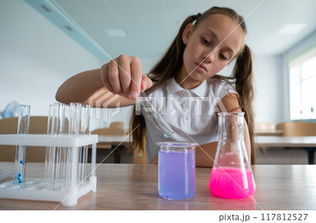 A schoolgirl conducts experiments in a chemistry lesson. Girl pouring colored liquids from a beaker. A schoolgirl conducts experiments in a chemistry lesson. Girl pouring colored liquids from a beaker. 117812527