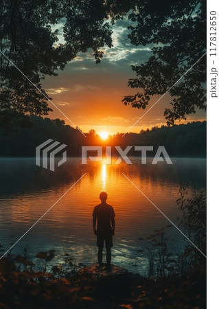 Silhouette of a Person Standing by a Tranquil Lake at Sunset Surrounded by Trees 117812650