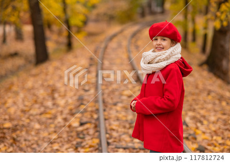 Caucasian girl in a red coat and beret walks along the railway tracks in the park in autumn. Caucasian girl in a red coat and beret walks along the railway tracks in the park in autumn. 117812724