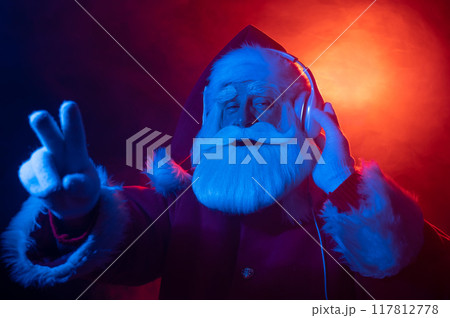 Santa Claus shows 2 fingers listening to music on headphones at a party. Blue red neon light and smoke. 117812778