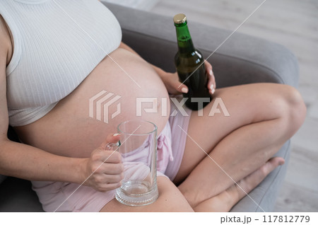 Faceless pregnant woman sitting on the sofa and holding a bottle of beer and a glass. Faceless pregnant woman sitting on the sofa and holding a bottle of beer and a glass. 117812779