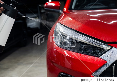 A man washes the headlights of a red car with a spray.  117812783