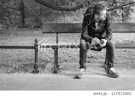 Lonely man sitting on the bench, monochrome. Tourist with backpack and adidas shoes thinking on bench in park, black and white. Active lifestyle. Alone guy in downtown. Lonely man sitting on the bench, monochrome. Tourist with backpack and adidas shoes thinking on bench in park, black and white. Active lifestyle. Alone guy in downtown. 117813002