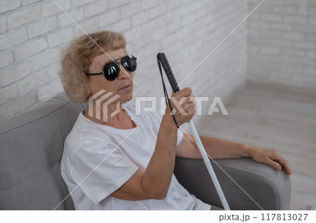 An elderly blind woman wearing sunglasses and with a tactile cane sits on the sofa. An elderly blind woman wearing sunglasses and with a tactile cane sits on the sofa. 117813027