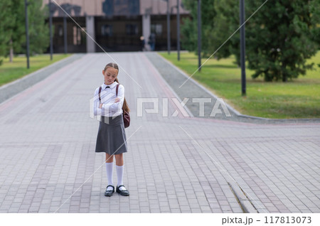 A sad caucasian girl in uniform and with a backpack holds her arms crossed on her chest stands outdoors after school. 117813073