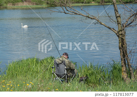 man with a fishing rod is sitting on the bank of the river and fishing. Stock photography. 117813077