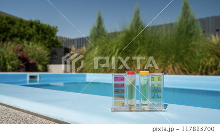 A pool water testing kit with pH and chlorine levels is placed on the pool's edge, with a soft-focus green backdrop. 117813700