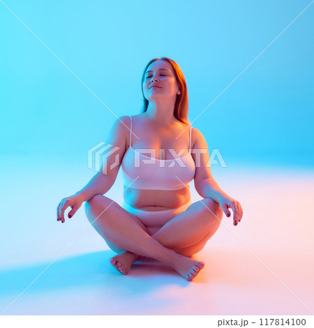 Portrait of young beautiful woman with chubby body shape sitting on floor and posing in underwear in relaxed pose against blue background background in neon light 117814100