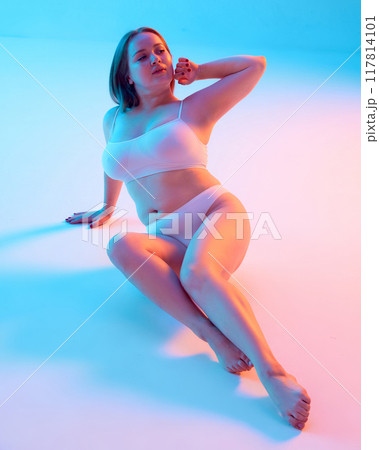 Portrait of young beautiful woman sitting confidently in textured swimwear and posing, showing self-love against blue background background in neon light 117814101