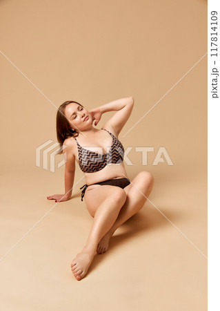 Portrait of young beautiful woman sitting confidently in textured swimwear and posing, showing self-love against beige studio background. 117814109