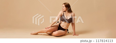 Portrait of attractive young woman with overweight body shape, posing in floor in black swimsuit against beige studio background. Portrait of attractive young woman with overweight body shape, posing in floor in black swimsuit against beige studio background. 117814113