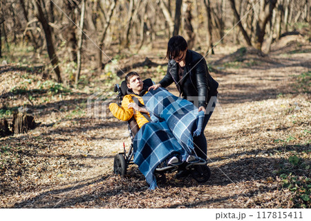 Mother adjusting blanket for child in wheelchair during outdoor walk in forest, caregiving, special needs support, family outings, disability inclusion. 117815411