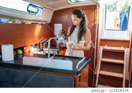Young woman standing and preparing tea in kitchen in cabin of modern yacht during sailing in sea. Yacht life concept 117816374