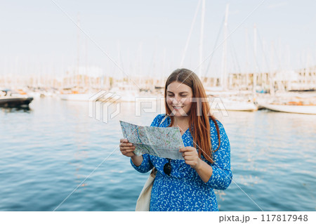 Vacation concept by exploring interesting places to travel. Beautiful Woman Searching locations at summer day. Woman with map walking along the waterfront, seaside. High quality photo 117817948