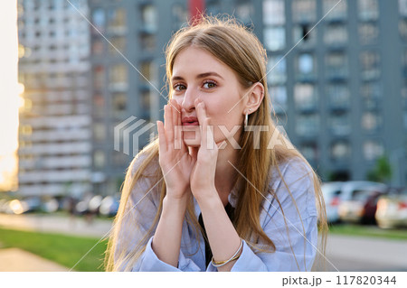 Young cheerful woman whispering secret, holding hands near mouth, outdoor 117820344
